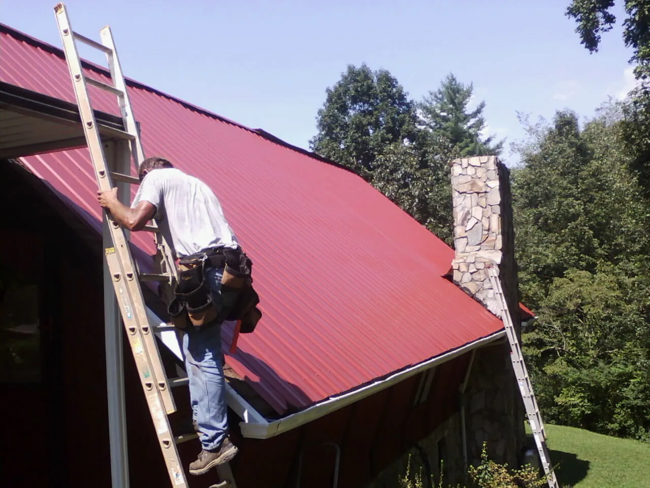Roofer on a ladder beside a red metal roof and stone chimney — Soffit & Fascia Repair by Roofing Brothers in Harford